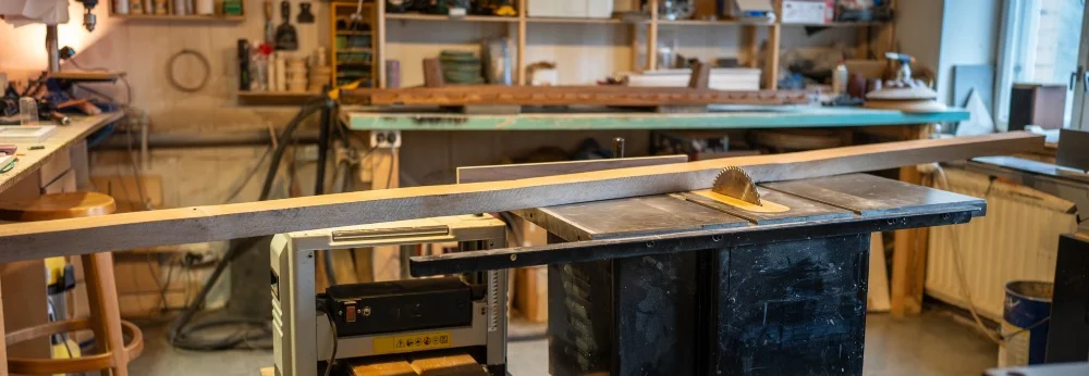 Tischkreissäge in einer Holzwerkstatt umgeben von Regalen mit Werkzeugen.